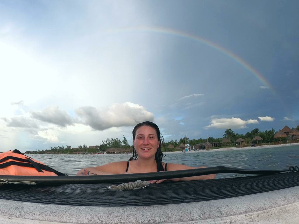 Solo female traveler smiling with a paddleboard and rainbow on Isla Holbox Mexico
