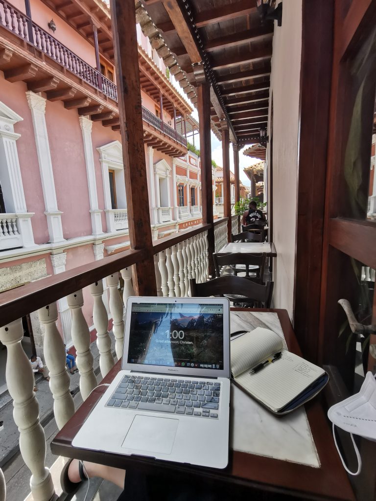 A digital nomad works remotely from her computer on a cafe terrace with pink colonial architecture in Cartagena Colombia