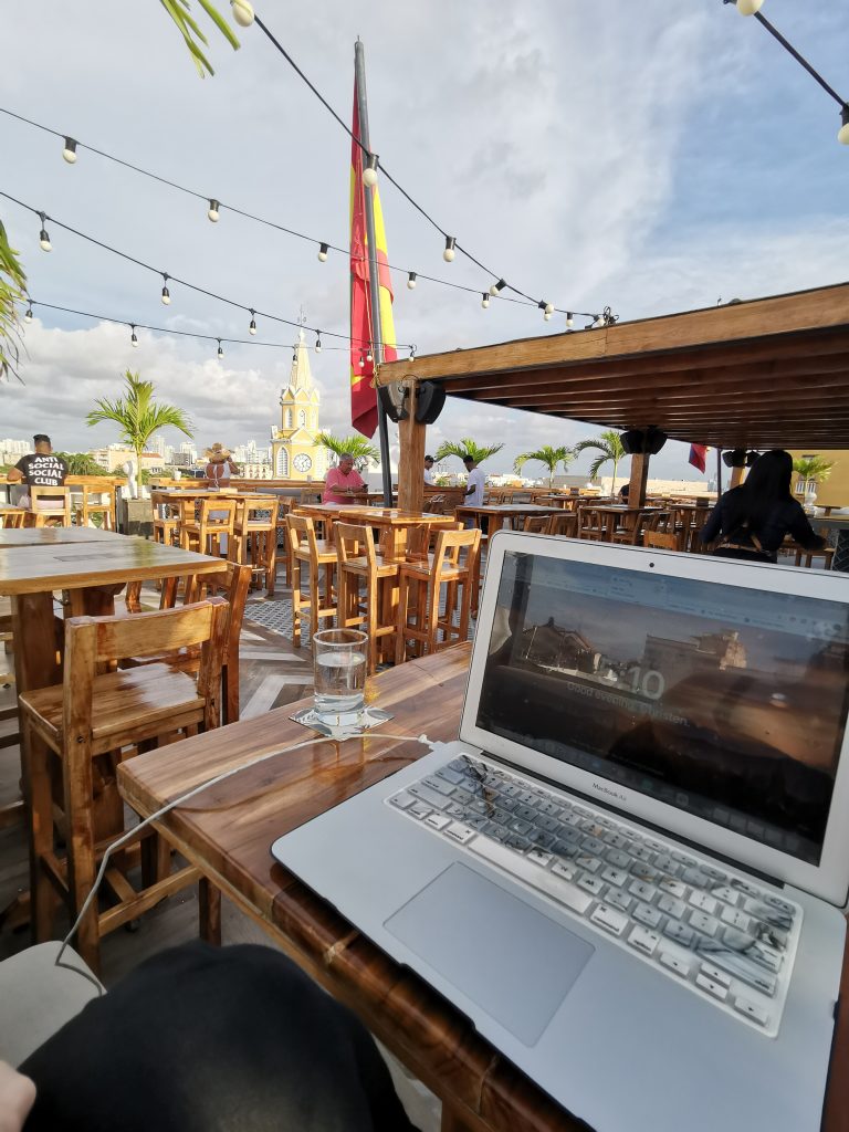 A digital nomad works from her computer from a rooftop bar in Cartagena Colombia with palm trees and the Colombian flag
